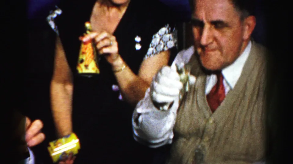 Albany 1950s home-movie frame of a man in a vest and tie raising a small glass while nearby guests hold colorful snack packages.