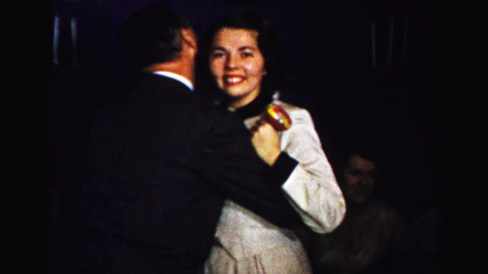 1950s Albany home-movie frame showing a smiling woman in a light dress dancing with a man in a dark suit at an indoor party.