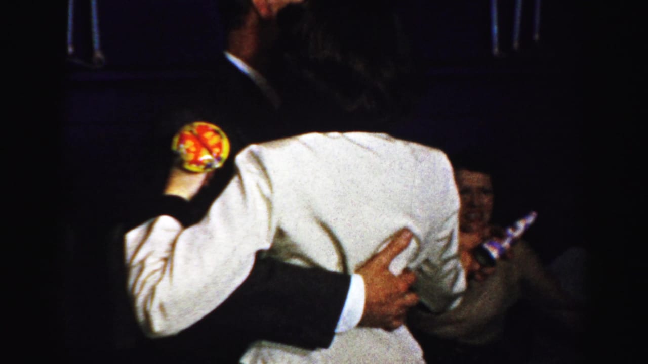 The couple continues dancing in a dark private interior, with the woman holding a colorful round party favor.