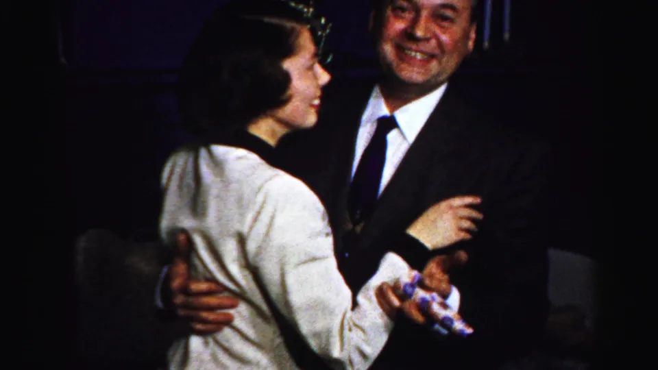 The woman turns in profile while dancing with a smiling suited man during the Albany indoor gathering.