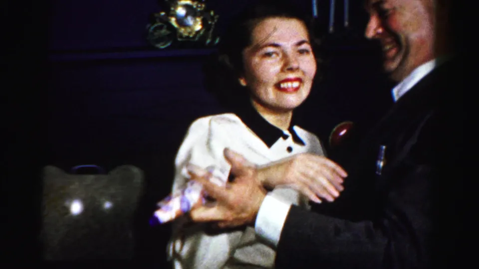 A smiling woman in a white dress dances with a suited man, with a small party favor visible in her hand.