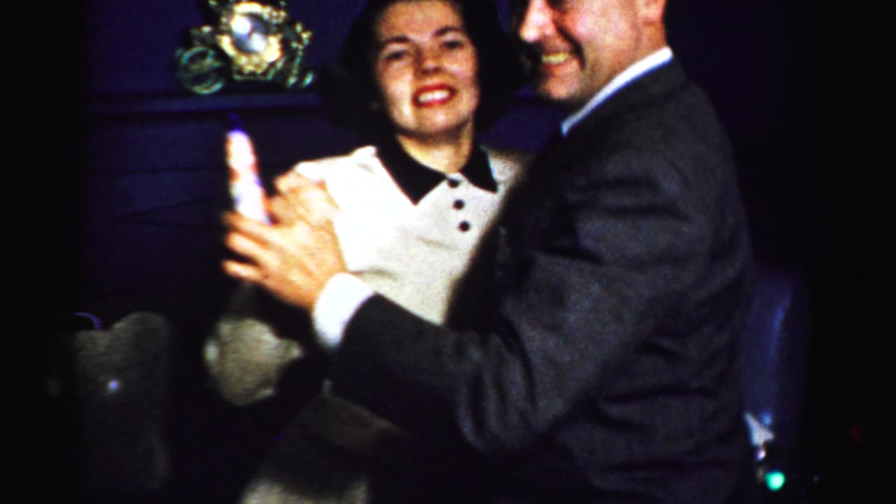 A smiling couple dances indoors in 1950s Albany footage, with the man in a dark suit holding a small glass.