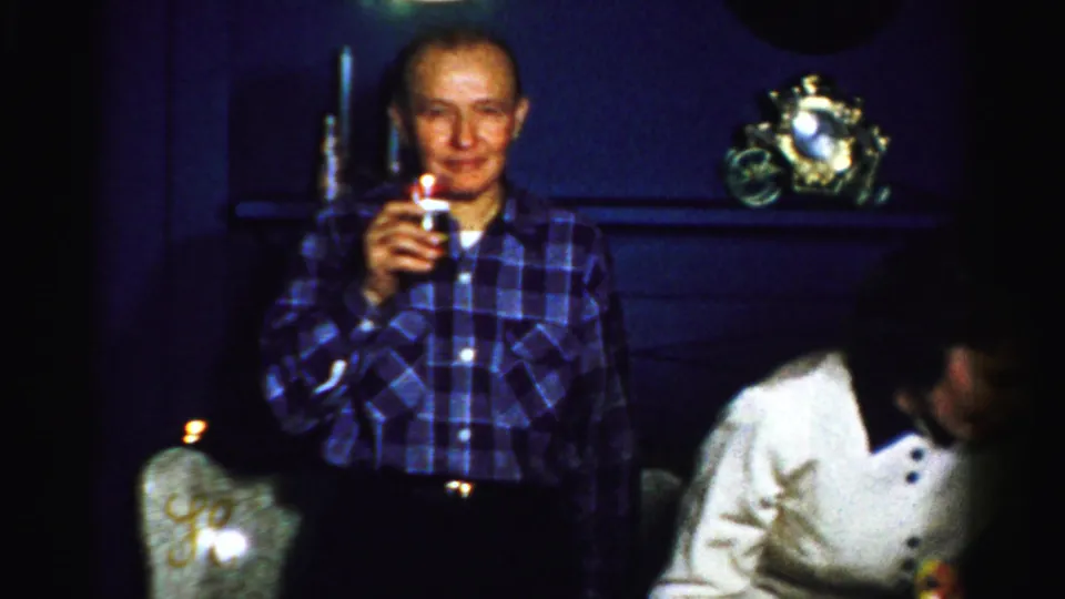 An older man in a blue plaid shirt raises a drink in a dim private interior, with a woman in a white sweater partly visible nearby.