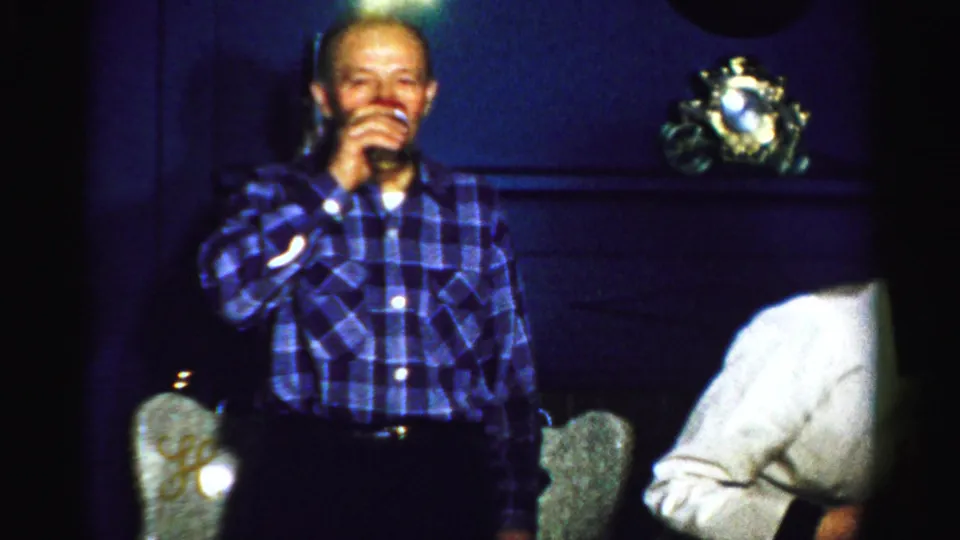 An older man in a blue plaid shirt drinks from a glass inside a dim Albany private interior, with chairs and a decorative clock behind him.