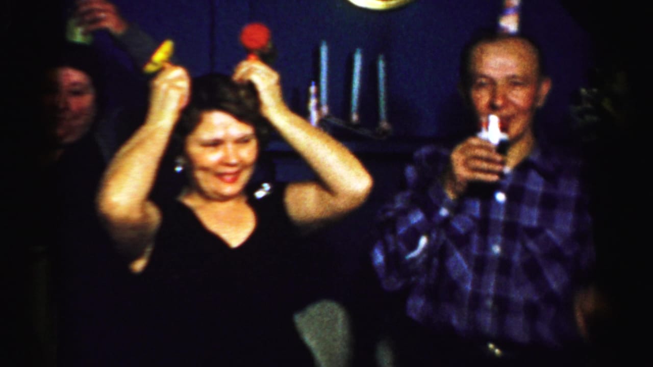 Woman in a black top raises two party noisemakers while adults stand nearby.