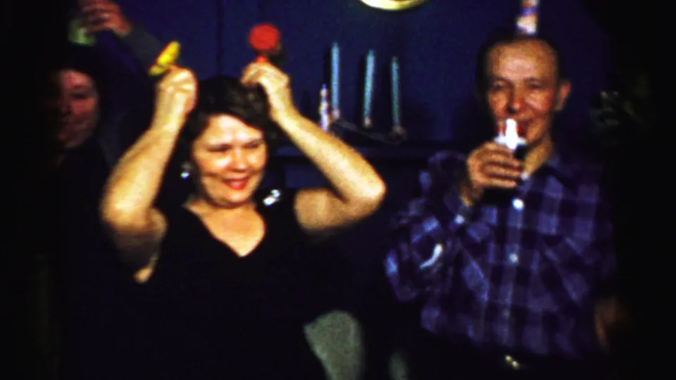 1956 Albany home-movie frame showing a woman in a black top lifting party noisemakers as other adults watch in a private room.