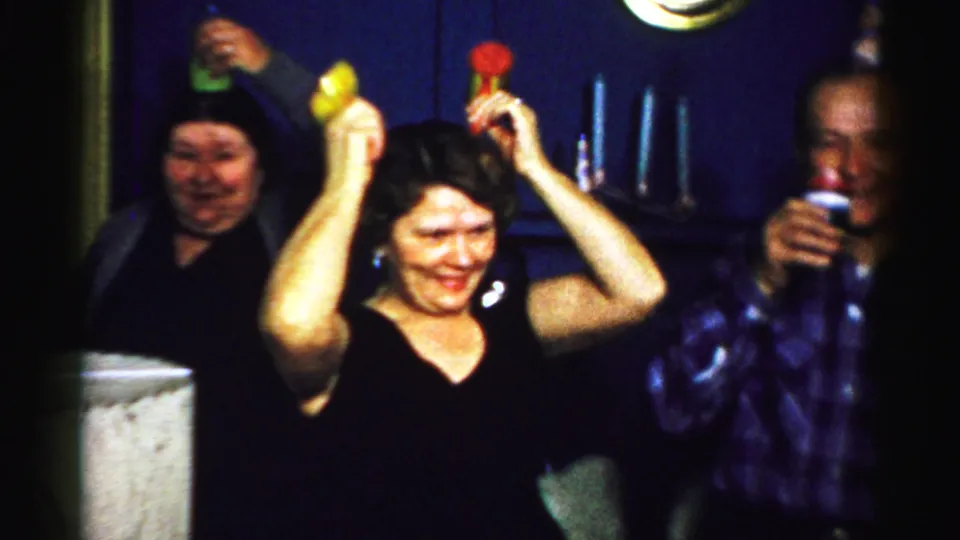 Albany 1956 indoor party frame with adults wearing paper hats, a woman lifting noisemakers, and a man partly visible with a drink.