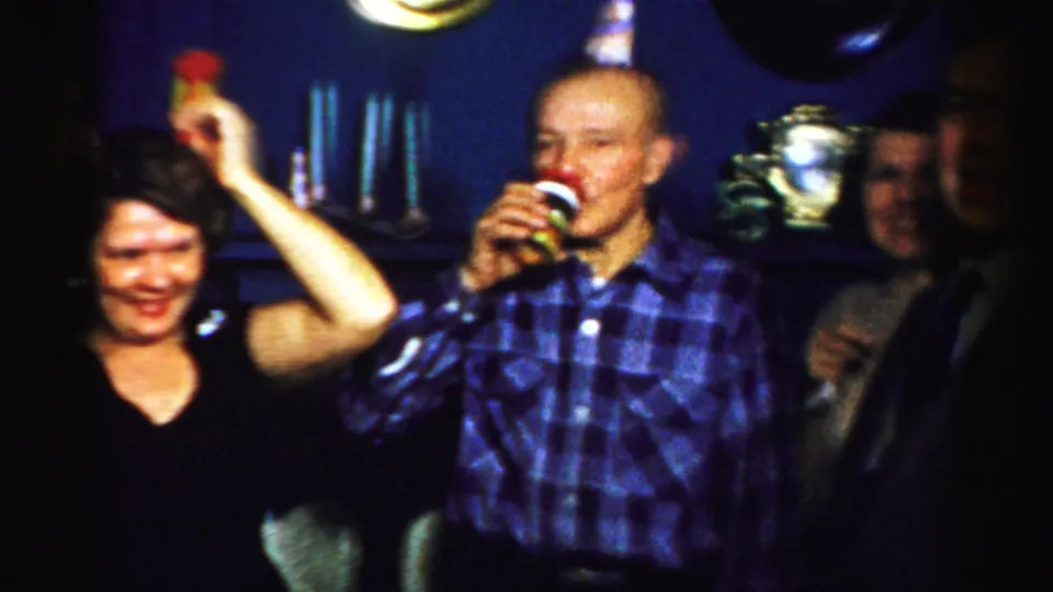 1956 Albany home-movie frame showing a man in a blue plaid shirt drinking and a woman beside him holding noisemakers in a private interior.