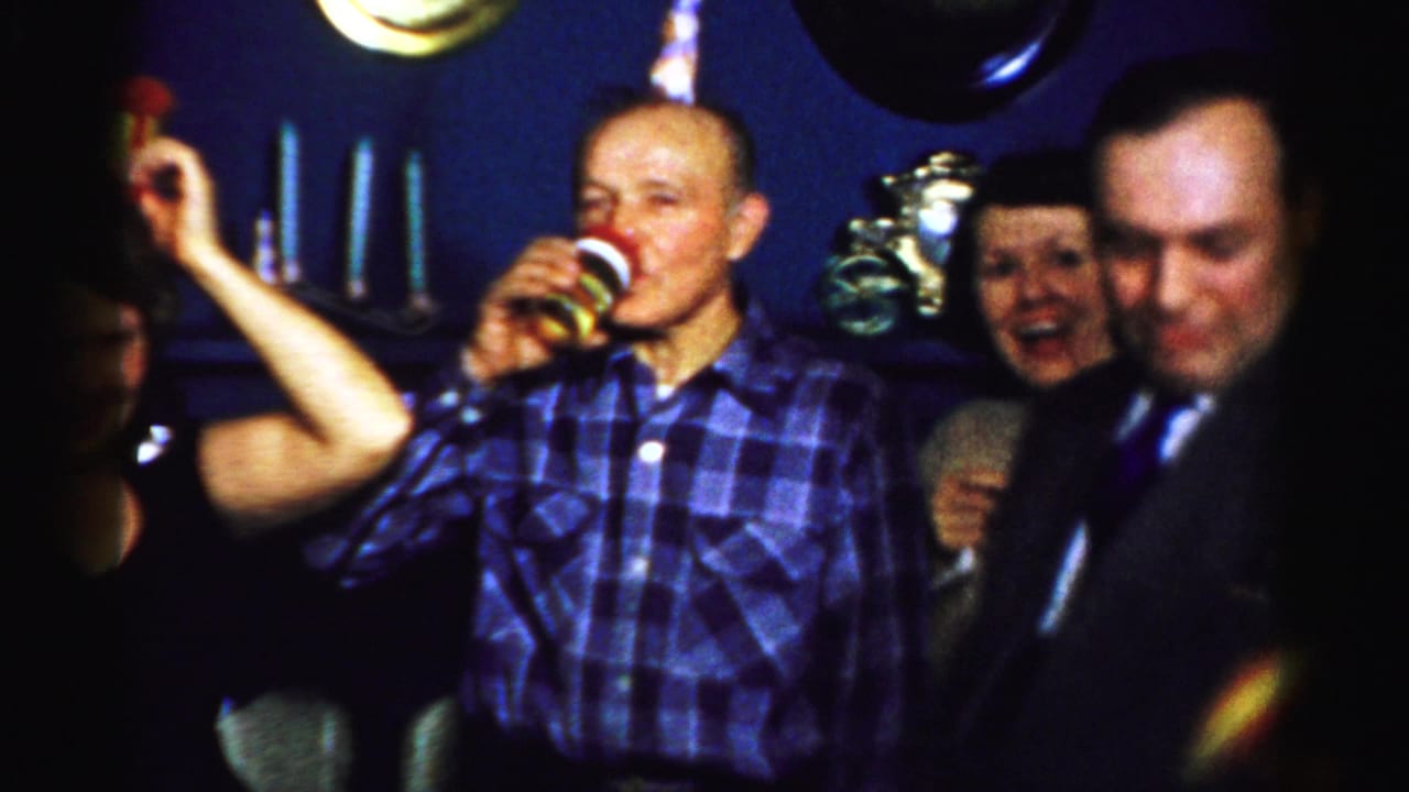 Man in a blue plaid shirt drinks from a cup while holding a party favor near other guests.