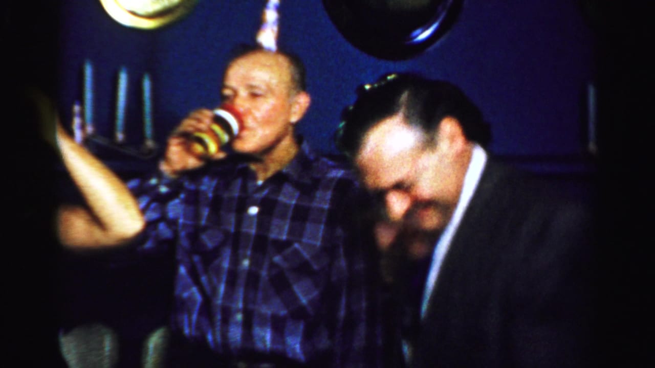 Man drinking from a cup as a suited guest leans into the foreground at an indoor party.