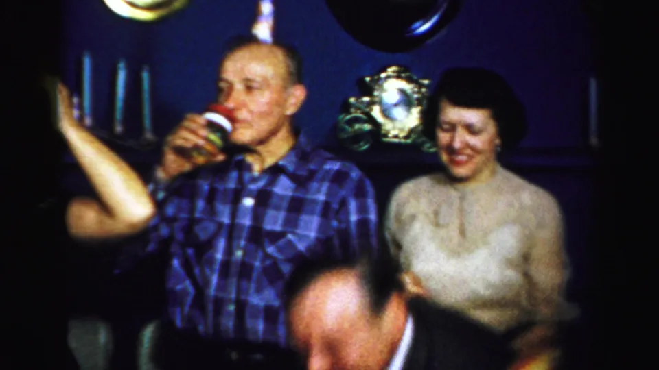A man in plaid stands with his cup beside a smiling woman in a light blouse during the 1950s Albany indoor celebration.