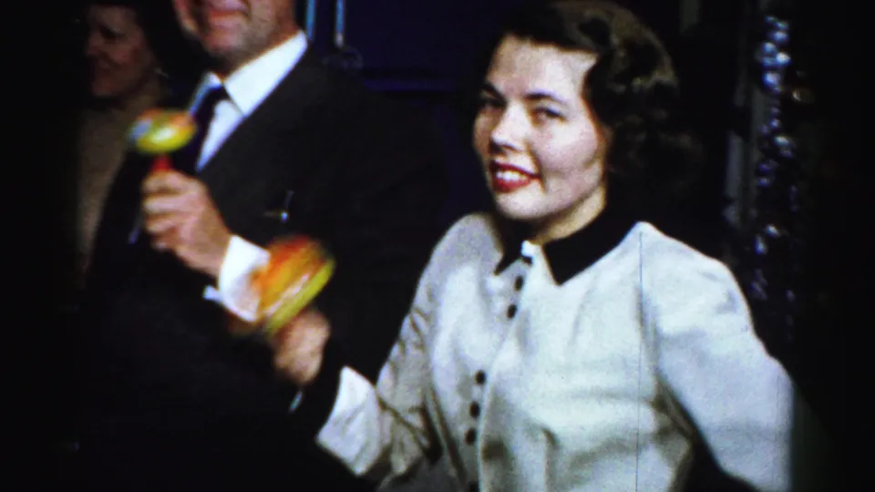A smiling woman in a light jacket holds maracas beside a suited man in the Albany home-movie party footage.