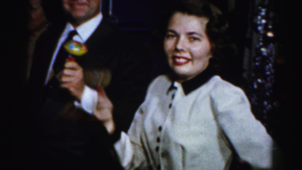 In 1956 Albany home-movie footage, a woman smiles at the camera as nearby adults hold layered party drinks in a dim interior.