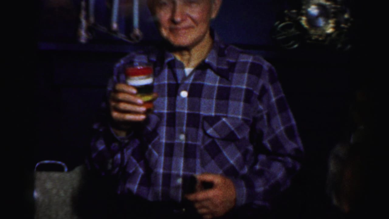 An older man in a blue plaid shirt holds a layered drink in a dim Albany party interior, with household decor behind him.