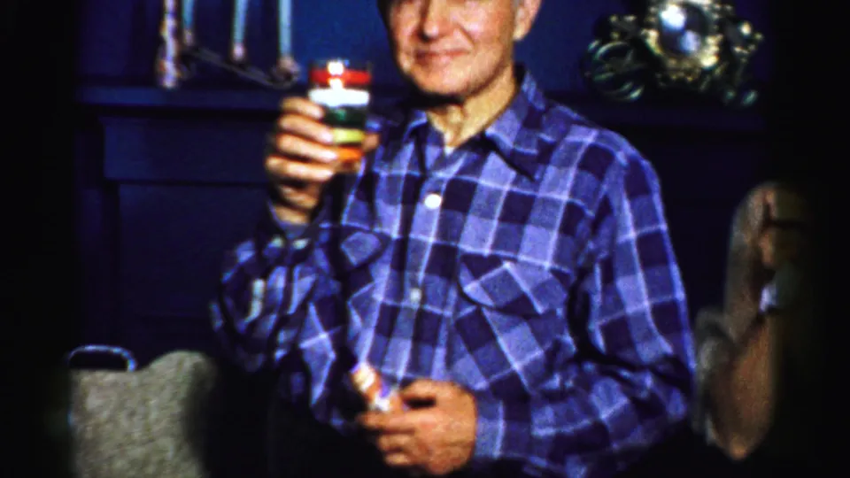 The older man stands in a plaid shirt and presents a layered drink toward the camera in 1950s Albany home-movie footage.