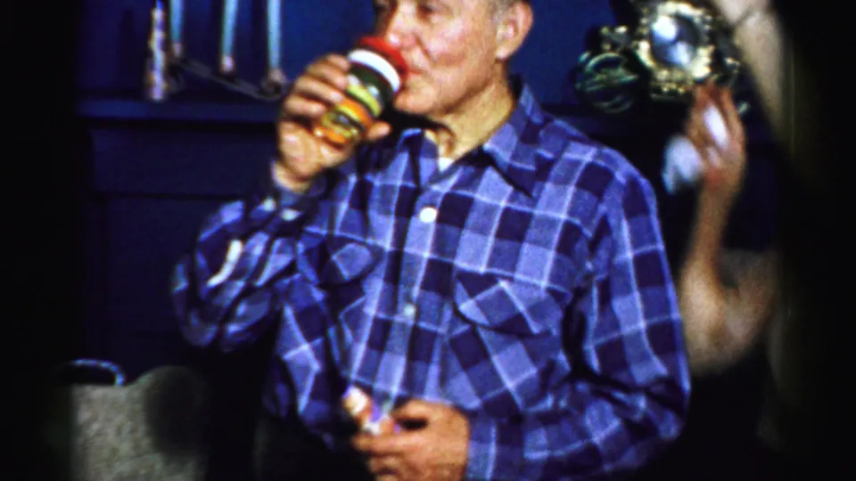 The older man drinks from a striped layered cup while another guest gestures beside him in the dim 1956 Albany party scene.