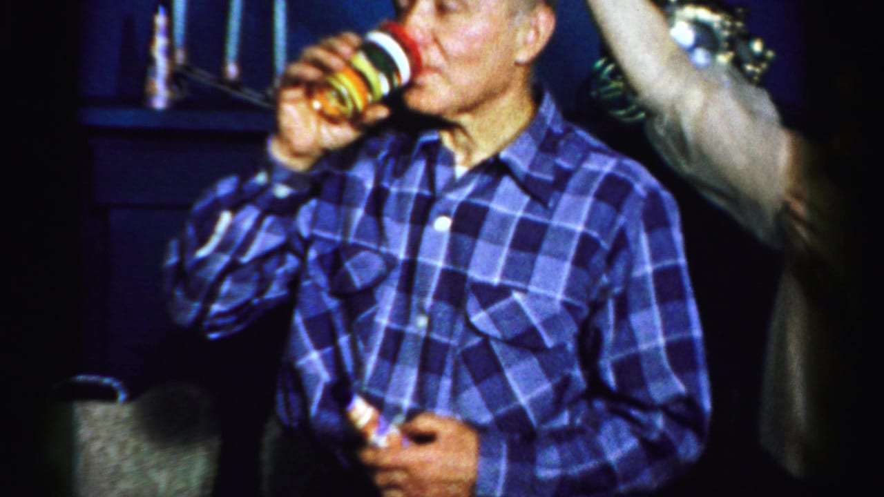 In this 1956 Albany home-movie frame, a man in a blue plaid shirt drinks from a colorful cup during an indoor party.