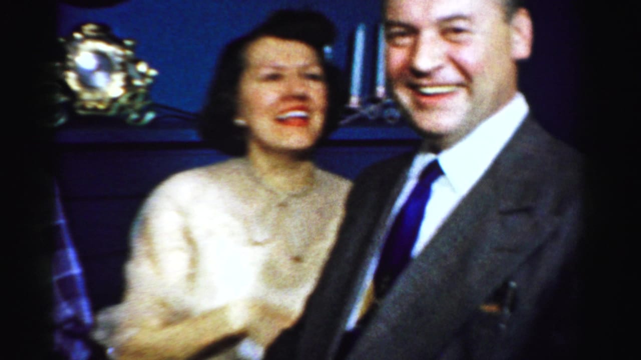 Smiling suited man stands beside a woman in a cream dress indoors.