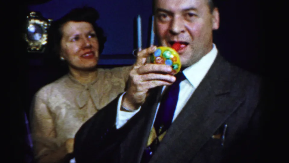 A man in a dark suit raises a bright party horn as a woman in a cream dress looks on inside the Albany party setting.