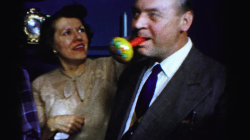 A suited man blows a yellow and red party horn while a woman smiles beside him in the 1956 Albany home-movie party scene.