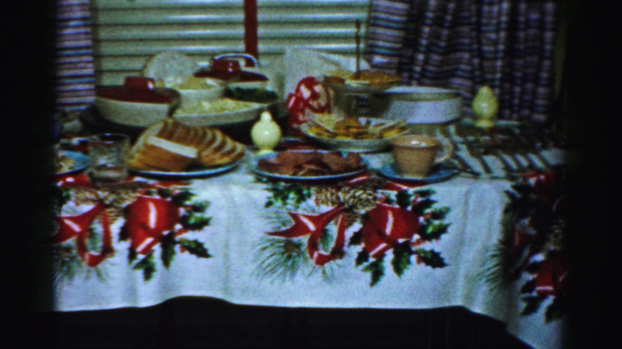 1956 Albany home-movie frame showing a holiday table set with bread, serving bowls, glasses, and a white cloth decorated with red bows and greenery.