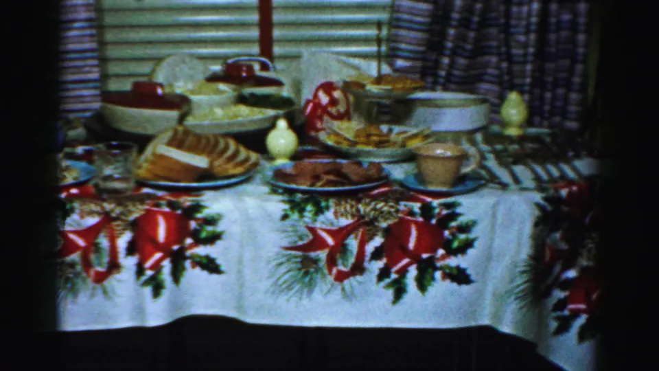 1956 Albany home-movie frame showing a holiday table set with bread, serving bowls, glasses, and a white cloth decorated with red bows and greenery.