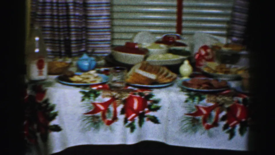 1956 Albany home-movie frame showing a holiday food table with a bottle, glass, bread, serving bowls, and decorated tablecloth.