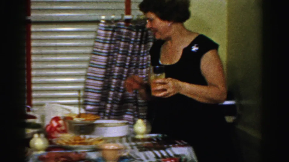 1956 Albany home-movie frame showing a woman in a black dress holding a drink beside a decorated holiday meal table.
