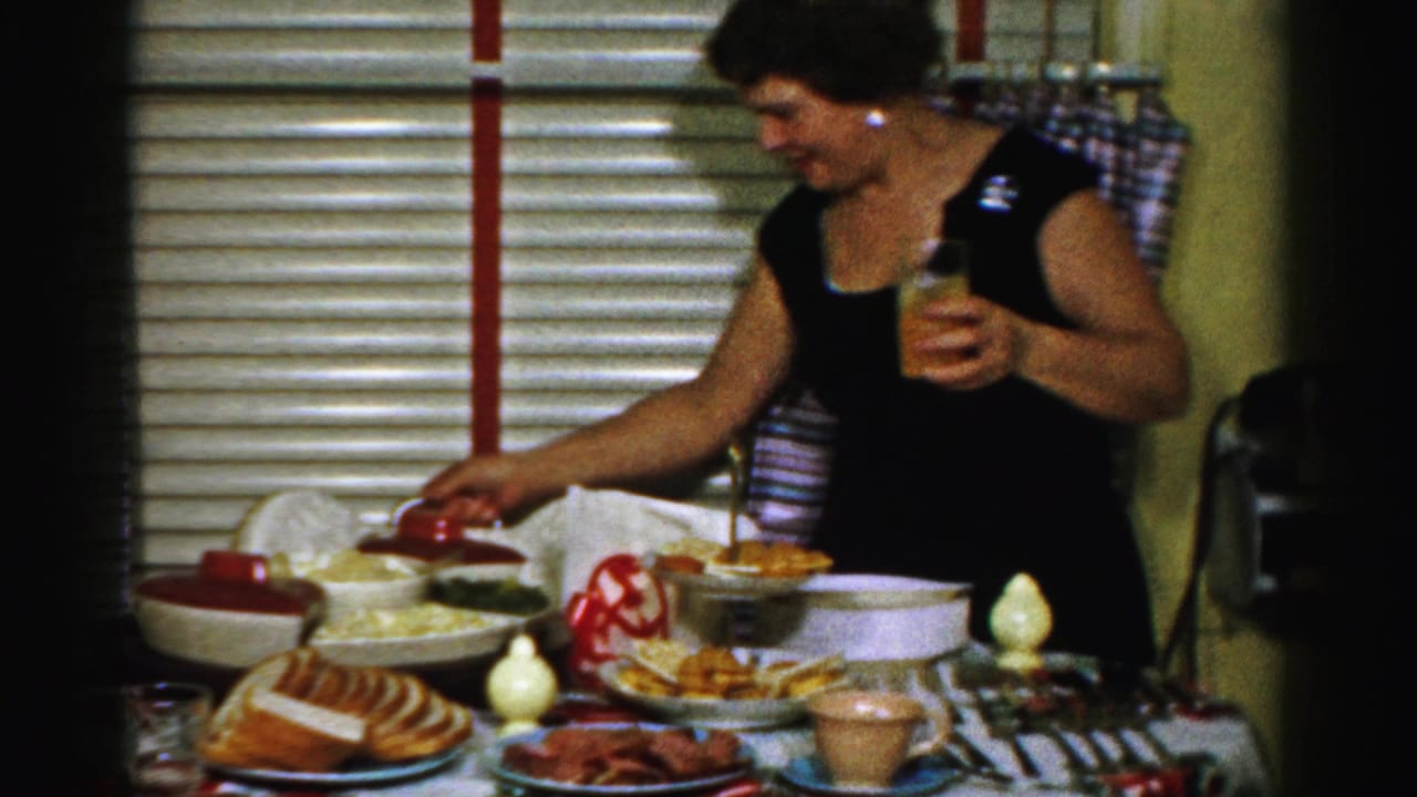 In Albany home-movie footage from 1956, a woman in a black dress reaches across a dinner table filled with dishes and bread.