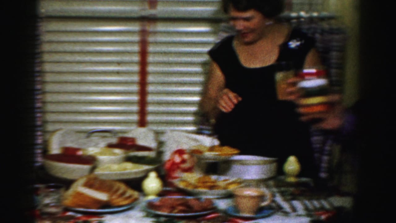 The woman stands by the dinner table with a glass of orange liquid as dishes, bread, and bowls fill the foreground.