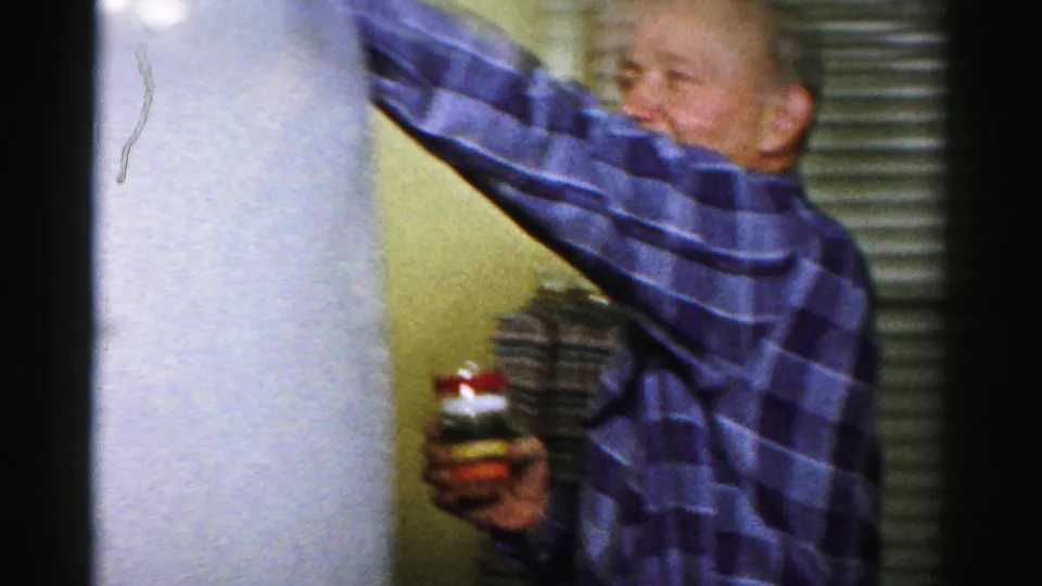 A man in a blue and purple plaid shirt raises one arm near a wall while holding a striped jar in the private interior.