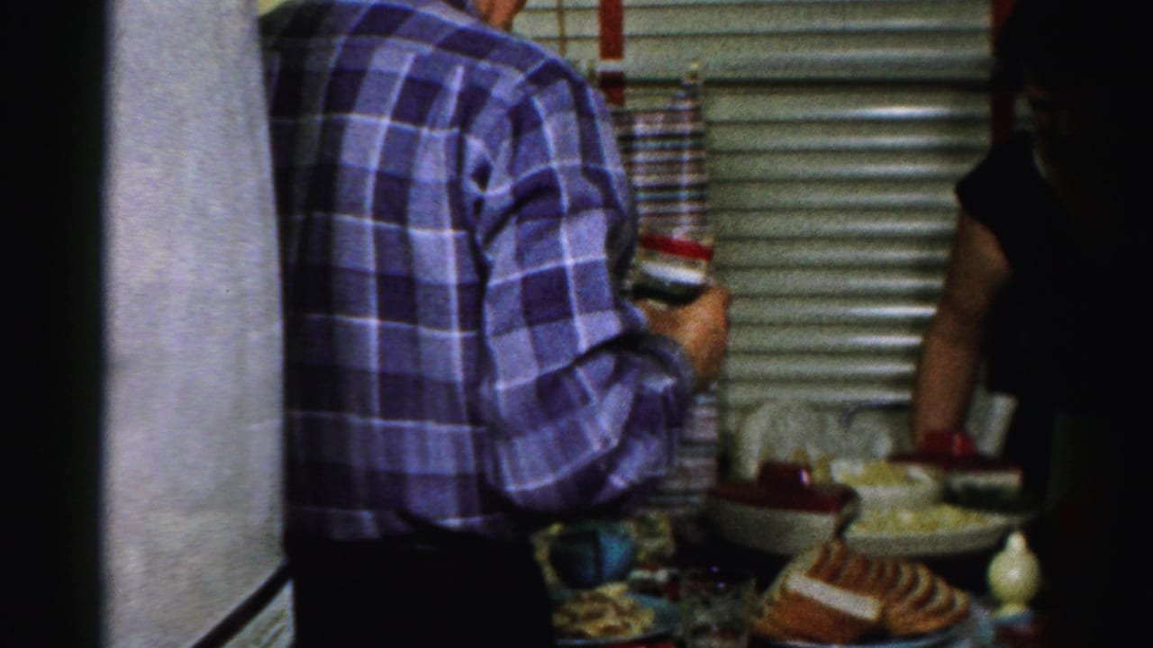 Man in plaid shirt stands beside a kitchen table covered with food.