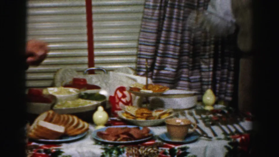 The kitchen table is set with sliced bread, meat, bowls, utensils, cups, and a patterned holiday cloth.