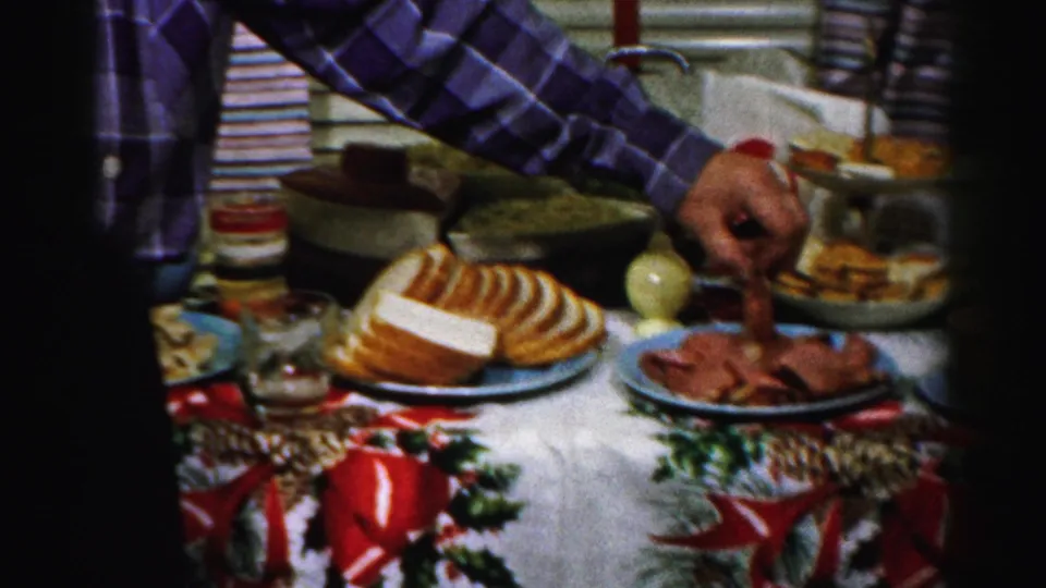 A person in a plaid shirt reaches over the Albany kitchen table to serve meat beside bread, bowls, and holiday table decorations.
