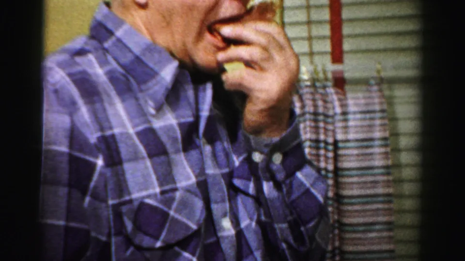 Adult man in a purple plaid shirt eats during a 1950s Albany holiday meal, with curtains and table setting nearby.