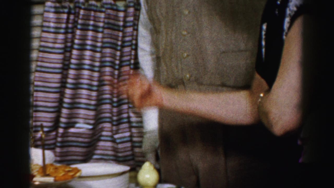Adults stand beside a party food table with crackers, dip, curtains, and a window blind.