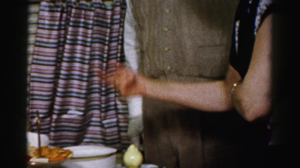 In this Albany home-movie frame, adults in mid-century clothing stand near a food table with crackers, dip, curtains, and a window blind.