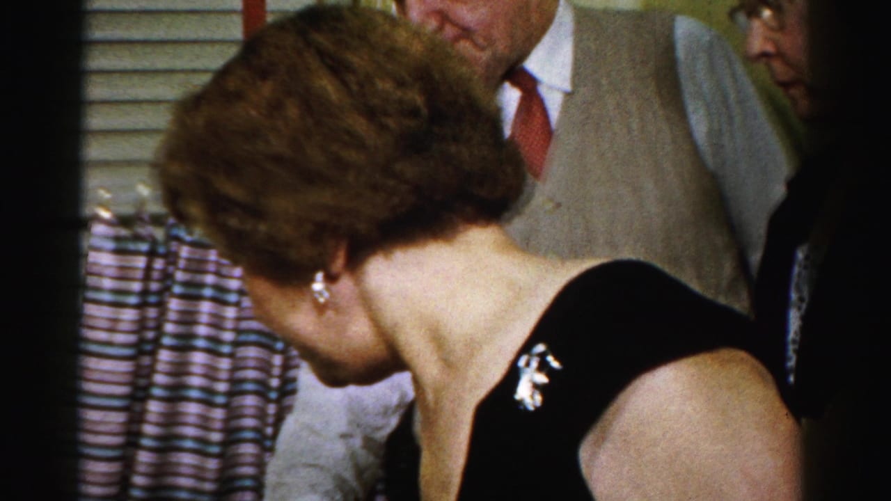 A woman in a black dress with a brooch leans toward the party table as adults stand behind her in a private Albany interior.