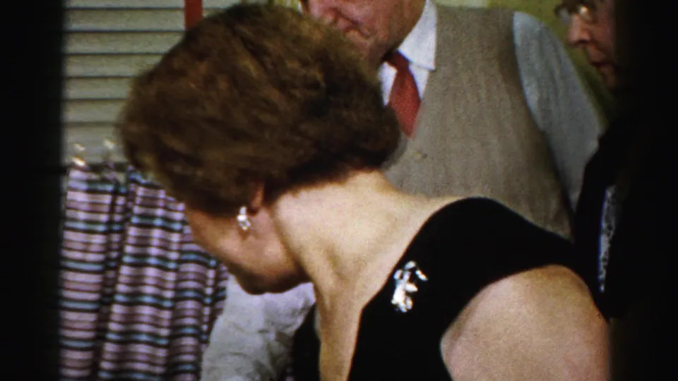 A woman in a black dress with a brooch leans toward the party table as adults stand behind her in a private Albany interior.