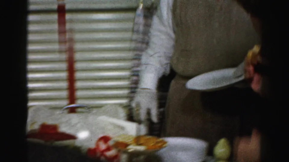 Adults serve themselves from a private party table with crackers, red dip, plates, plaid curtains, and window blinds visible.
