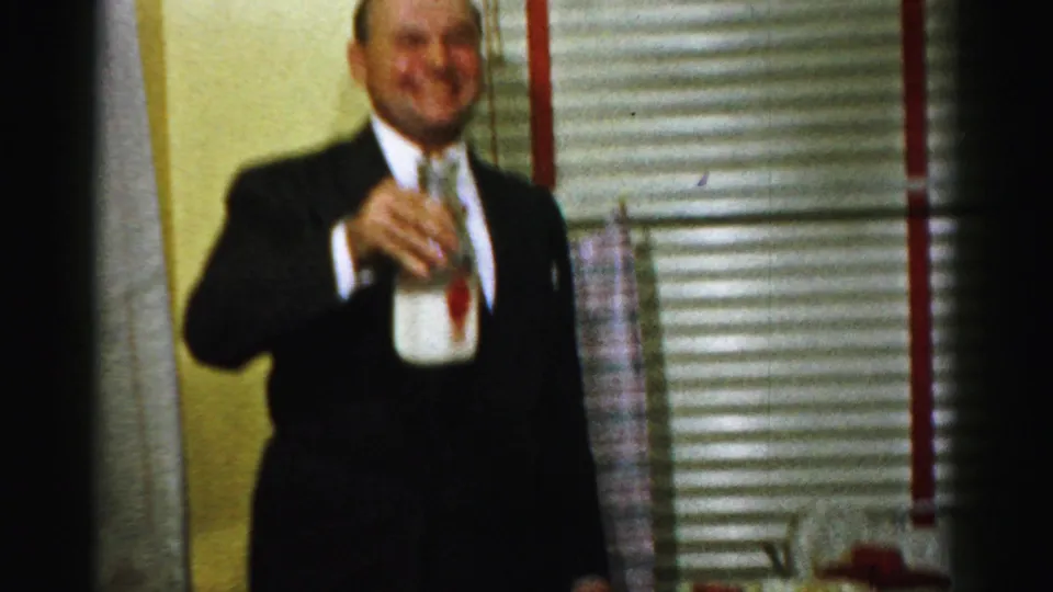 A man in a dark suit holds a bottle near the food table in a 1950s Albany private-party home-movie frame.