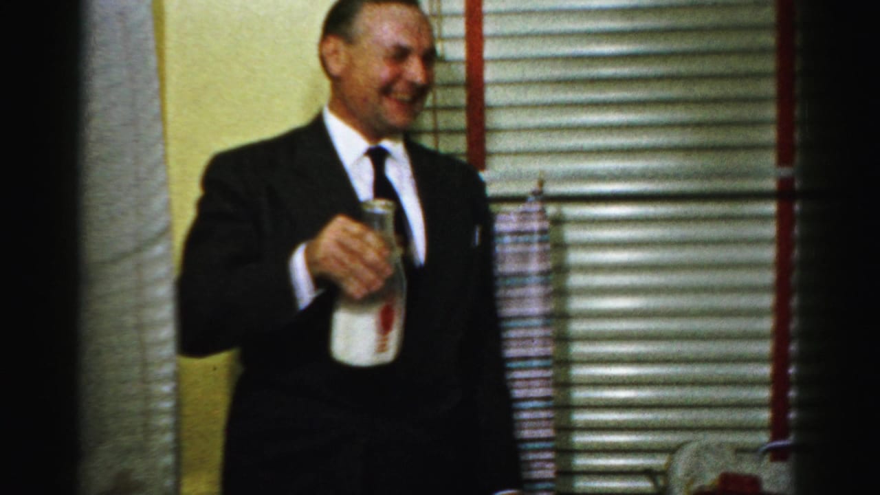In Albany home-movie footage from 1956, a suited man smiles indoors while holding a glass milk bottle near venetian blinds.