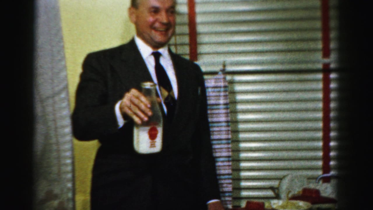 Smiling man in suit holds a milk bottle while standing near blinds.