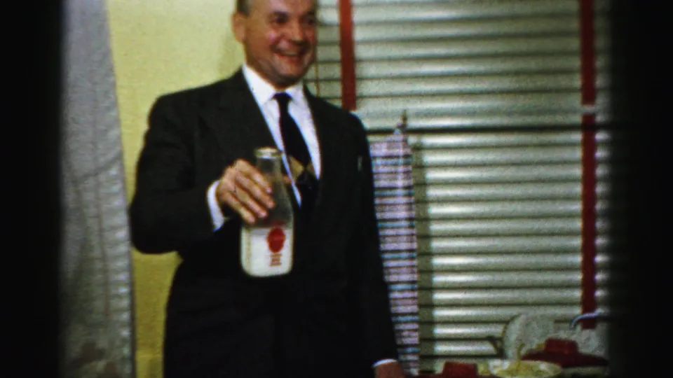 The suited man continues smiling with the glass milk bottle raised in a 1956 Albany kitchen scene.