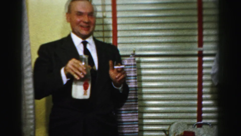 A suited man in the Albany kitchen holds a glass milk bottle and appears to gesture while holding a cigarette.
