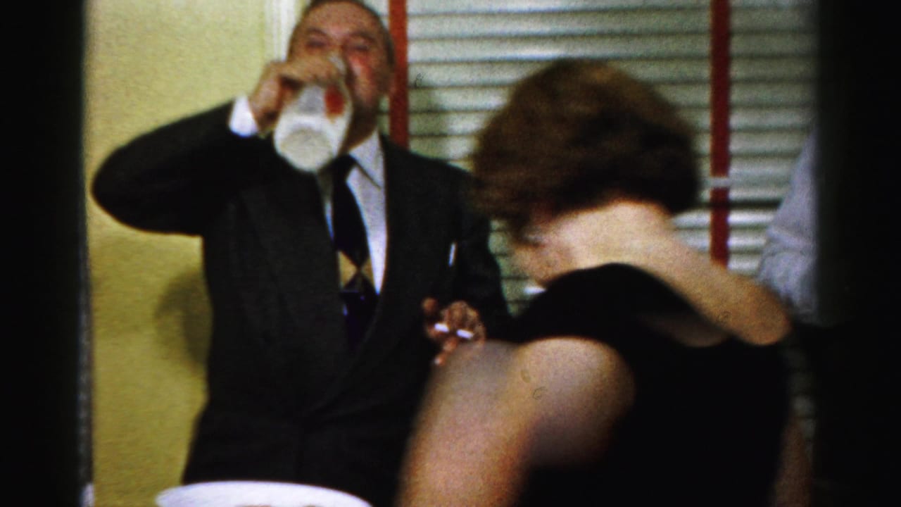 In this Albany 1950s office party frame, a suited man drinks from a glass as a woman in a black dress moves in front of him.