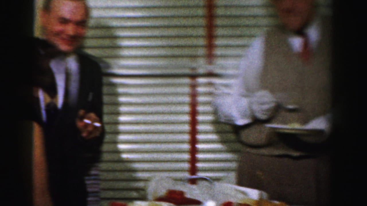 Adults stand near blinds during a party, with one man smiling and another holding a serving tray.