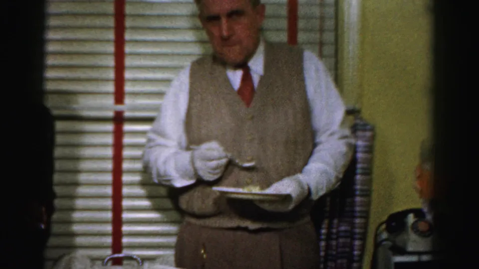 A waiter in a vest, tie, and white gloves holds a plate or tray inside the 1950s Albany office party setting.