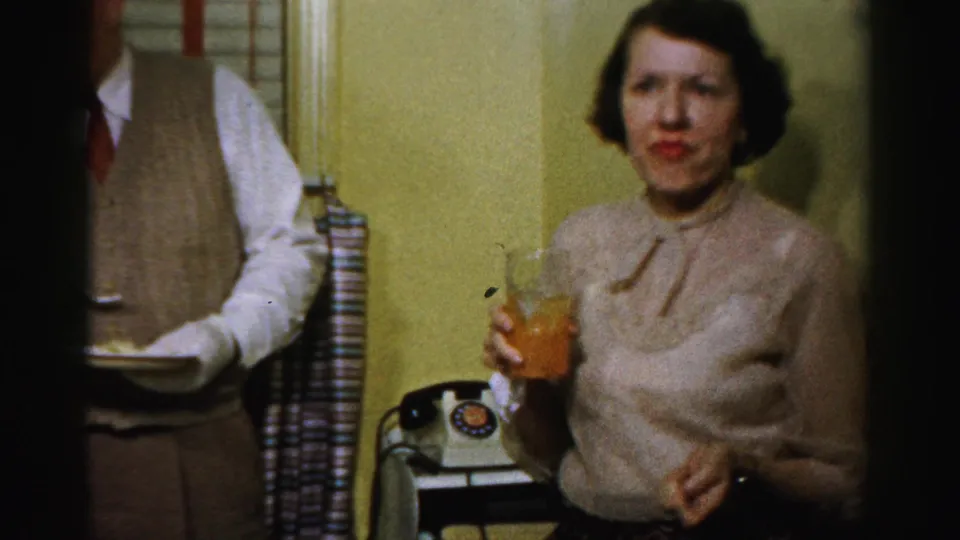 A woman in a light bow-collar sweater holds a drink near a rotary telephone during the Albany 1950s office party.
