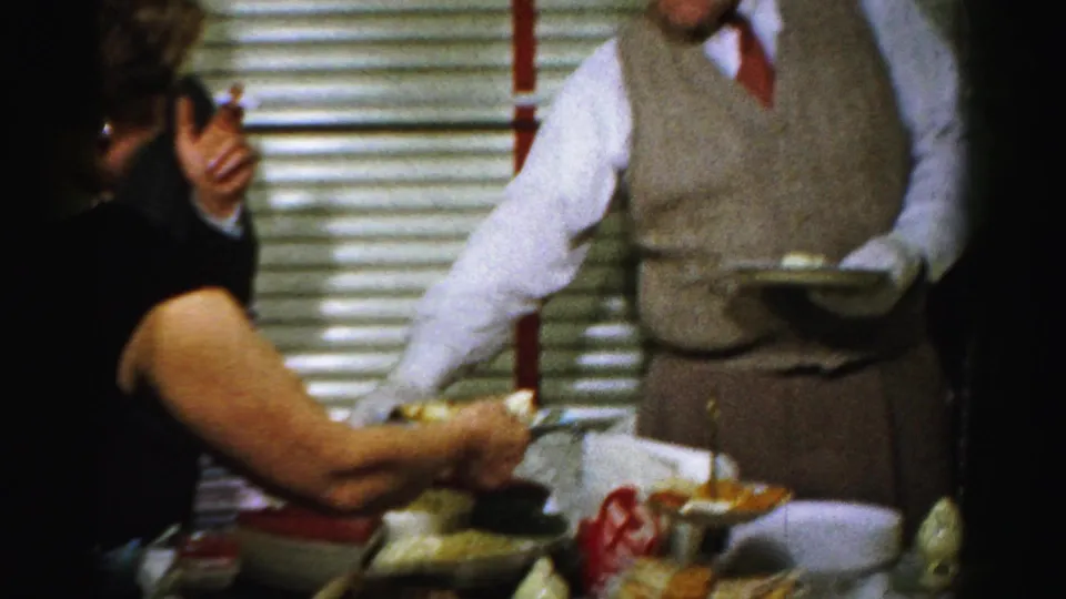 A man in a sweater vest and tie offers or holds a plate while guests gather around a 1950s Albany buffet.